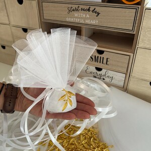 May include: White tulle favor bags with gold olive branch charms. The bags are tied with white satin ribbon and are filled with white satin pouches. The pouches are decorated with gold olive branch charms. The bags are arranged on a white surface. The text "Start each day with a grateful heart" and "Smile Everyday" is visible on the wooden drawers in the background.
