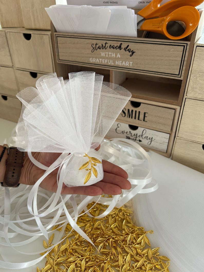 May include: White tulle favor bags with gold olive branch charms. The bags are tied with white satin ribbon and are filled with white satin pouches. The pouches are decorated with gold olive branch charms. The bags are arranged on a white surface. The text "Start each day with a grateful heart" and "Smile Everyday" is visible on the wooden drawers in the background.