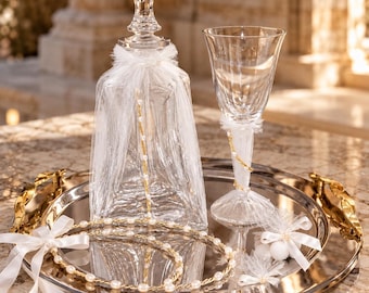 May include: A clear glass decanter and a matching stemmed glass, both adorned with white tulle and gold-toned pearl strands. They are arranged on a round silver-toned metal tray with gold-toned handles, alongside a circular pearl-accented wreath.