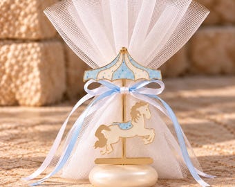 May include: A carousel-themed favor decoration featuring a gold-tone metal horse and a blue and white polka-dot canopy. The ornament is set on a round, cream-colored base and is wrapped in a white tulle fabric tied with a light blue ribbon.