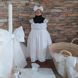 May include: A white christening dress with lace trim, a white candle with a bow, a white basket with a lace trim, a white towel, and a small bottle with a white bow.