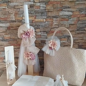 Può includere: Un set di regali per il battesimo che include una candela bianca con decorazioni floreali rosa e bianche, un asciugamano bianco con bordino in pizzo, una borsa beige con un abbellimento floreale rosa e una piccola bottiglia di liquido trasparente con un fiocco rosa.