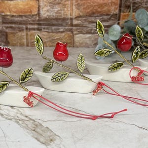 May include: Three ceramic rose decorations. Each rose has a red bud, green leaves, and a gold stem. They sit in white boat-shaped dishes. Red strings with the year "2026" are attached. The background is a mix of brick and marble.