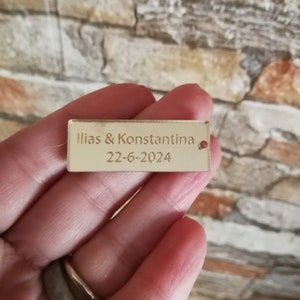 May include: A gold-colored acrylic tag with the text "Ilias & Konstantina" and the date "22-6-2024" engraved on it.