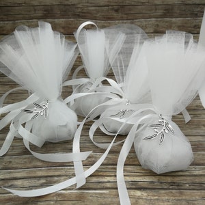 May include: Three white tulle favor bags with white satin ribbon ties and silver olive branch charms. The bags are on a wooden surface.