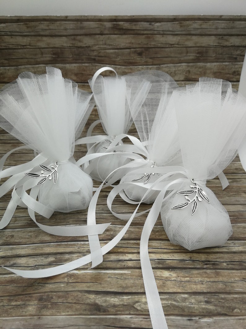 May include: Three white tulle favor bags with white satin ribbon ties and silver olive branch charms. The bags are on a wooden surface.