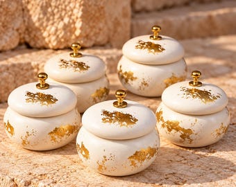 May include: A set of four round, off-white ceramic jars with lids, each featuring a gold metallic knob handle and irregular gold leaf accents scattered across the surface of the lids and the rounded bodies of the containers.
