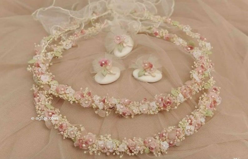 May include: Two floral headbands and three matching hair accessories. The headbands and accessories are adorned with small pink and white flowers, pearls, and green leaves. The accessories are on white oval bases. The background is a soft pink fabric.