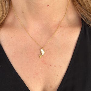 May include: A gold-colored necklace featuring a dolphin pendant. The dolphin has a white body and gold-colored accents. The necklace is worn around the neck and hangs down.