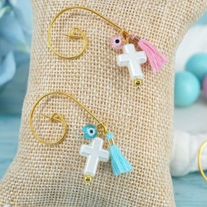 May include: Two gold-toned ear cuffs with a spiral design. Each cuff features a white cross charm, a small tassel, and an evil eye bead. One cuff has a pink tassel, the other a blue tassel. The background includes blue and pink candies.