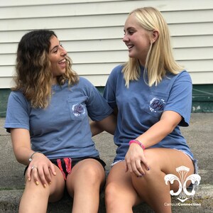 May include: Two people wearing blue t-shirts with white embroidery. The text on the shirts reads "Big" and "Little".