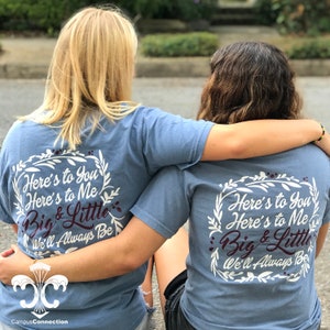May include: Two people wearing light blue t-shirts with a white floral wreath design and the text "Here's to You Here's to Me Big & Little We'll Always Be" printed on the back. The person on the left is wearing a black skirt and the person on the right is wearing black shorts.