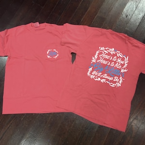 May include: Two coral pink t-shirts with a white pocket on the left shirt and a white graphic on the right shirt. The graphic says "Here's to You Here's to Me Big & Little We'll Always Be".