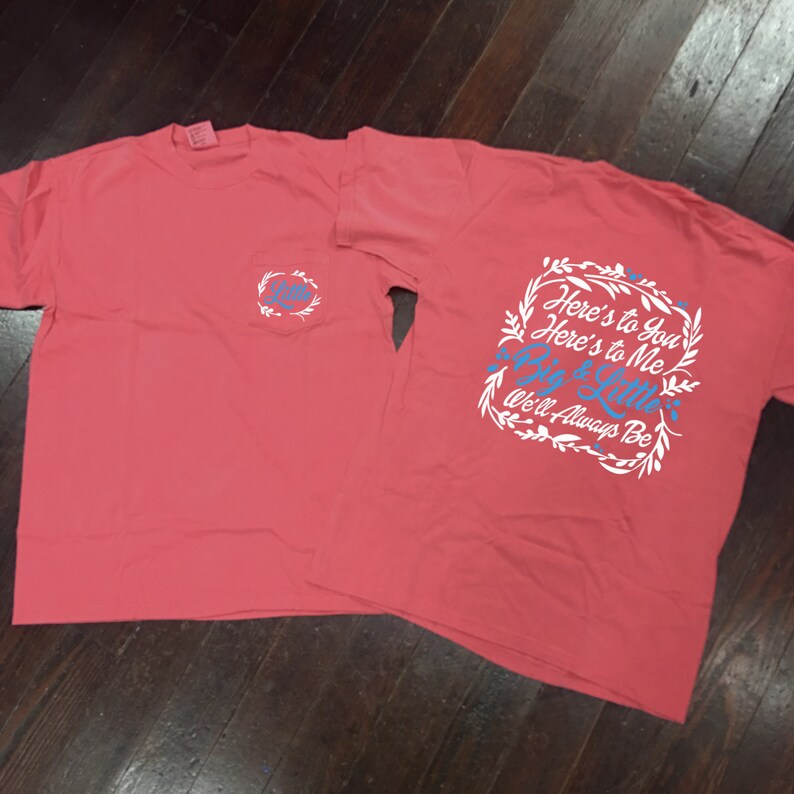 May include: Two coral pink t-shirts with a white pocket on the left shirt and a white graphic on the right shirt. The graphic says "Here's to You Here's to Me Big & Little We'll Always Be".