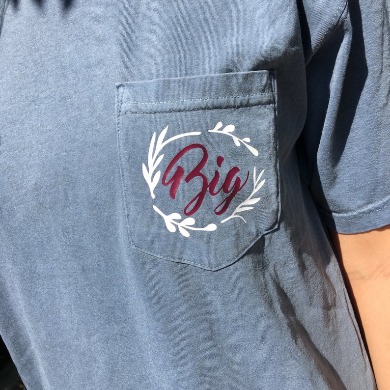 May include: A blue t-shirt with a white pocket featuring a white floral wreath with the word "Big" in burgundy lettering.