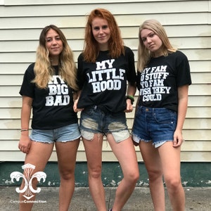 May include: Three women wearing black t-shirts with white text. The text on the shirts reads "My Big Bad", "My Little Hood", and "My Fam O Stuff Yo Fam Wish They Could". The women are wearing denim shorts and standing in front of a white wooden wall.