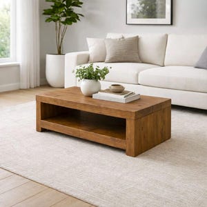 May include: A rectangular, wooden coffee table with a lower shelf. The table is a warm brown colour and sits on a light-coloured rug. A white sofa and a potted plant are in the background. A white vase with greenery sits on the table.