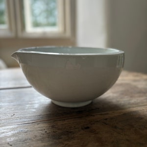 May include: A white ceramic mixing bowl with a pouring spout. The bowl has a rounded shape and sits on a small, circular base. The interior is a slightly lighter shade of white. The bowl is on a wooden surface.
