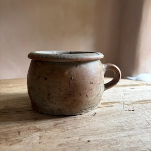Peut inclure: Un petit pot en céramique de couleur beige, de forme ronde, doté d'un bord épais et saillant ainsi que d'une anse latérale unique. La surface présente une texture tachetée avec des points brun foncé et un aspect terreux.