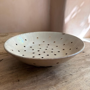 Könnte beinhalten: Eine flache, runde Keramikschale mit einer glänzenden, cremeweißen Glasur und einer Reihe gleichmäßig verteilter Abflusslöcher am Boden und an den Seiten. Die Schale hat einen glatten, geschwungenen Rand und ein leicht unregelmäßiges Aussehen.