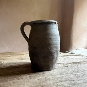 May include: A cylindrical stoneware jug with a textured, matte grey finish and a single side handle. The exterior surface features horizontal ridges and small, irregular indentations throughout the body, with a flat rim and a slightly tapered base.