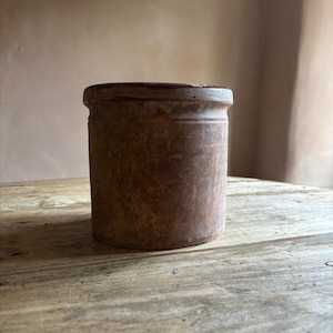 Puede incluir: Un jarrón cilíndrico de cerámica marrón con un borde ligeramente ensanchado. El jarrón tiene un aspecto envejecido, lo que sugiere edad y uso. Se encuentra sobre una superficie de madera rústica, con un fondo neutro.