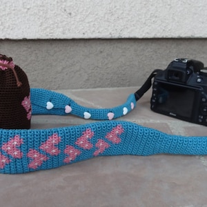 May include: A blue crocheted camera strap with pink hearts and white beads. A brown crocheted pouch with pink hearts is also included.