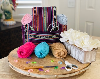 May include: A colorful woven yarn bag with a pocket, filled with pink and gray yarn balls. Other yarn balls in pink, blue, and tan are on a wooden surface, along with a crochet hook, scissors, and stitch markers. A decorative floral arrangement is also present.
