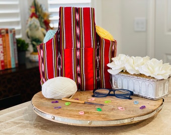 May include: A red and white striped fabric yarn tote bag with a yellow handle. The bag is filled with yarn balls and is sitting on a wooden table with a white ball of yarn, knitting needles, and stitch markers.
