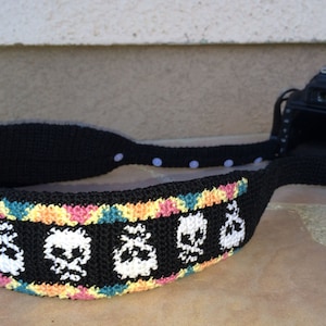 May include: Black crochet camera strap with a repeating pattern of white skulls. The strap has a colorful border with pink, yellow, and teal accents. The strap is attached to a black DSLR camera.