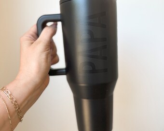 Könnte beinhalten: Ein schwarzer isolierter Becher mit Henkel und Deckel. Das Wort "PAPA" ist auf der Seite eingeprägt. Der Becher wird vor einem weißen Hintergrund gehalten.