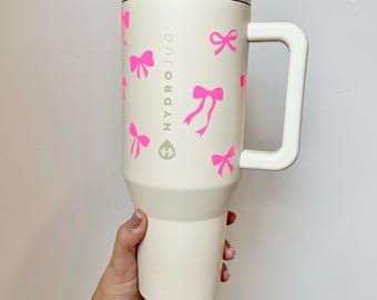 Könnte beinhalten: Eine weiße Reise-Thermoskanne mit einem rosa Schleifenmuster und dem Text "HYDROJUG" an der Seite.