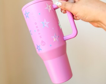 Könnte beinhalten: Ein pinkfarbener Hydrojug-Becher mit Griff und Deckel. Der Becher ist mit holografischen Sternaufklebern verziert. Der Text "HYDROJUG" ist auf der Seite aufgedruckt. Der Becher wird von einer Person gehalten.