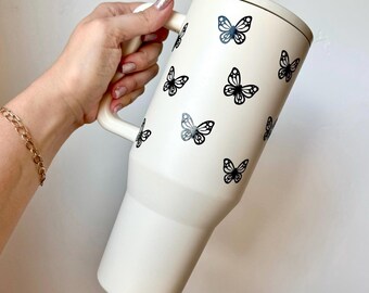 Könnte beinhalten: Eine weiße Reise-Tasse mit einem schwarzen Schmetterlingsmuster. Die Tasse hat einen Griff und einen Deckel.