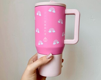 Könnte beinhalten: Rosa Reisebecher mit einem weißen Regenbogenmuster und dem Text "HYDROJUS" an der Seite. Der Becher hat einen Griff und einen Deckel.