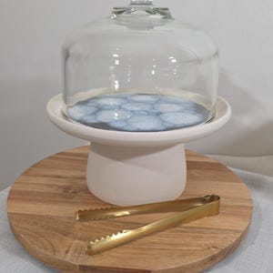 Può includere: Una campana di vetro trasparente con pomello poggia su un piedistallo bianco con base a fantasia blu e bianca. Un tagliere in legno e pinze color oro completano la presentazione. Ideale per esporre prodotti da forno o piccoli oggetti.