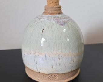 May include: A handcrafted ceramic bottle with a cork stopper. The bottle has a rounded shape with a light green and white glaze. The base and the neck are a natural, unglazed clay color. The cork stopper is a light brown color.
