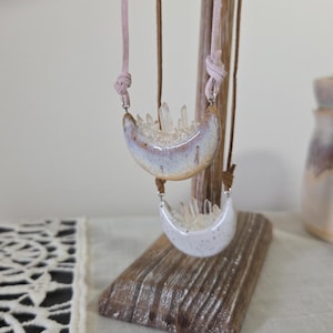 May include: Two crescent-shaped necklaces with crystal accents hang from a wooden display. The top necklace has a light brown and white glaze, while the bottom one is white. Both are suspended by pink and brown cords.