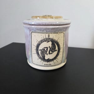 May include: A ceramic jar with a lid, featuring a Pisces zodiac design. The jar has a lavender and cream color scheme, with a label displaying two fish and the word "PISCES". The jar is on a black surface.