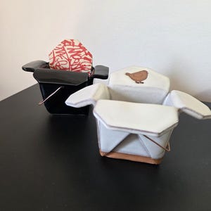 Puede incluir: Dos recipientes de cerámica para llevar. Uno es negro y contiene una forma abstracta roja y blanca. El otro es blanco con un diseño de pájaro marrón y una base marrón. Ambos tienen asas de alambre de cobre.