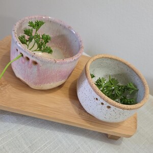 Può includere: Due ciotole in ceramica fatte a mano, una rosa e una bianca, sono su un vassoio di legno. La ciotola rosa ha una glassa macchiata e piccoli fori. Entrambe le ciotole contengono rametti di prezzemolo verde. Ideali per salse.