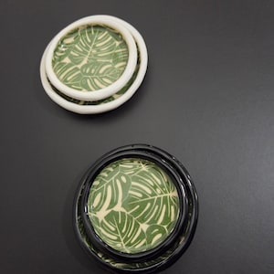 May include: Two decorative ceramic dishes. One set has a white rim and the other has a black rim. Both have a green and cream-colored leaf pattern on the inside. The dishes are arranged on a dark gray surface.