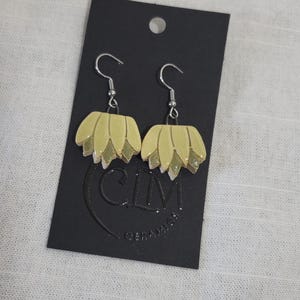 May include: A pair of ceramic earrings with a flower design. The earrings are a light yellow color with green accents. The earrings are hanging from silver hooks.