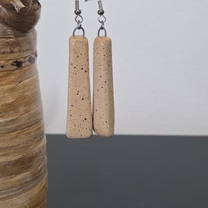 May include: A pair of handmade, beige ceramic earrings with a speckled pattern. The earrings are rectangular with rounded edges and hang from silver-colored ear wires. The earrings are displayed against a neutral background.