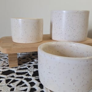 May include: Three speckled ceramic containers in varying sizes. The containers are off-white with small brown speckles. They are arranged on a small wooden tray with a bamboo base. The background is a neutral color.