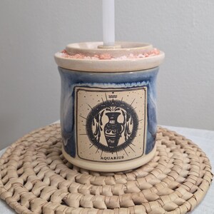 May include: A blue and cream ceramic candle holder with an Aquarius zodiac sign label. The candle holder has a white candle inserted in the top and sits on a woven mat.