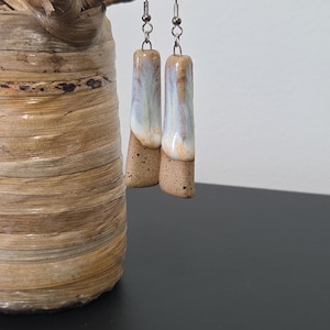 Könnte beinhalten: Ein Paar handgefertigte Keramikohrringe. Jeder Ohrring hat eine rechteckige Form mit einem gesprenkelten, beigefarbenen Sockel und einer glasierten Oberseite mit Weiß-, Blau- und Brauntönen. Die Ohrringe hängen an silberfarbenen Haken.