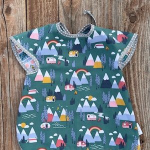 May include: A green bib with a camping-themed pattern featuring mountains, rainbows, and camper vans. The bib has short sleeves and a gray trim with white polka dots. A tie at the neck secures the bib.