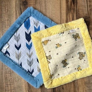 May include: Two baby blankets with different patterns. One blanket has a blue faux fur border and a white background with blue and gray arrows. The other blanket has a yellow faux fur border and a white background with a pattern of bears and honeycombs.