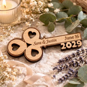 May include: Wooden key-shaped wedding favor with the names "Sara & Justin" and the date "June 16 2029" engraved on it. The key has heart-shaped cutouts and a dark brown border. The background includes flowers and a lit candle.